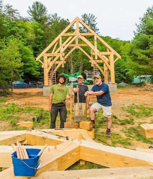 Students stand in front of a timber frame structure they are building in a workshop