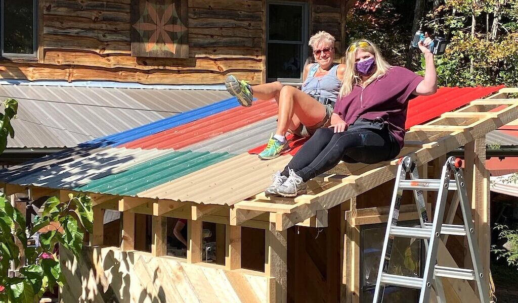 Two women laugh and pose on top of tiny house during women's advanced carpentry class