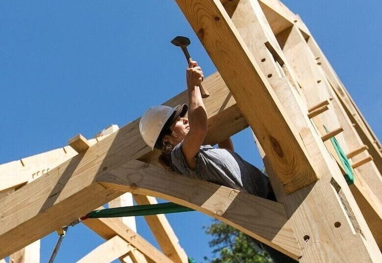 A female student wearing a safey helmet and harness uses a mallet while learning to build in a timber frame workshop