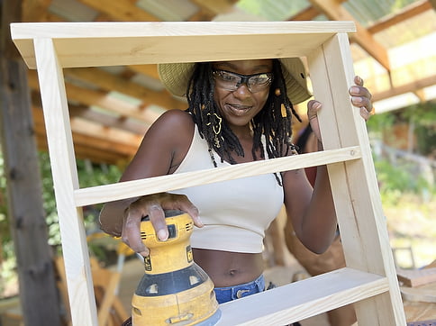 Black woman using a palm sander to sand the bookshelf she made in a basic carpentry class