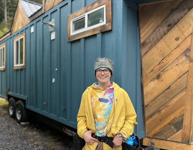 Person in raincoat and tool belt stands outside tiny house during workshop