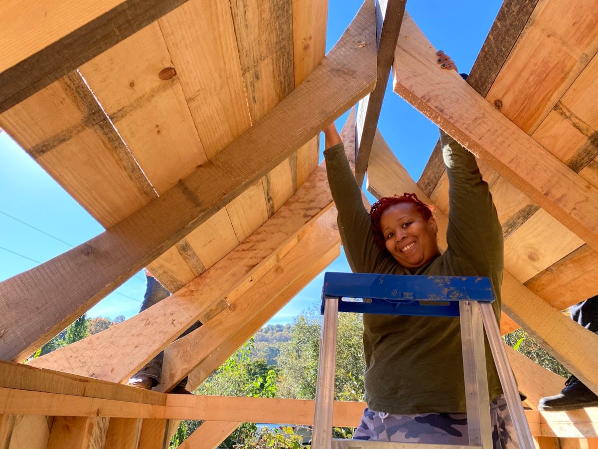 A woman smiles down as she is reacuhing up to the rafters while working on a Tiny House self-build project