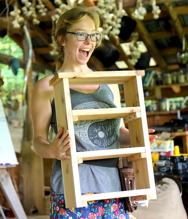 Woman poses excitedly with her final shelf project after women's basic carpentry class
