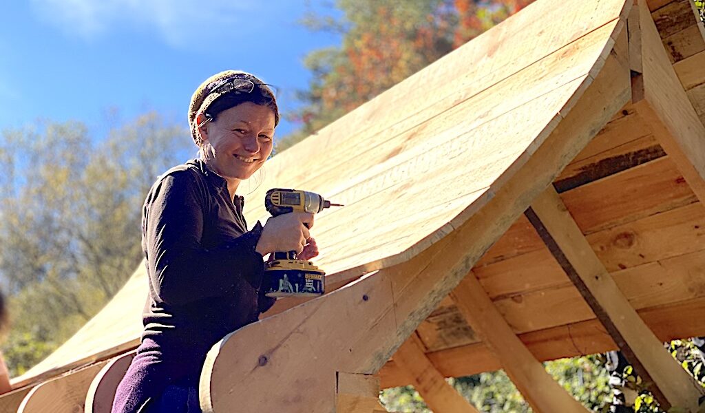 Student poses on ladder using drill on roof during advanced carpentry class