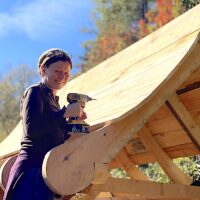 Student poses on ladder using drill on roof during advanced carpentry class