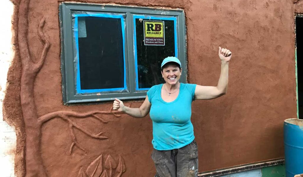 woman in a blue shirt standing in front of a tiny house with natural plaster
