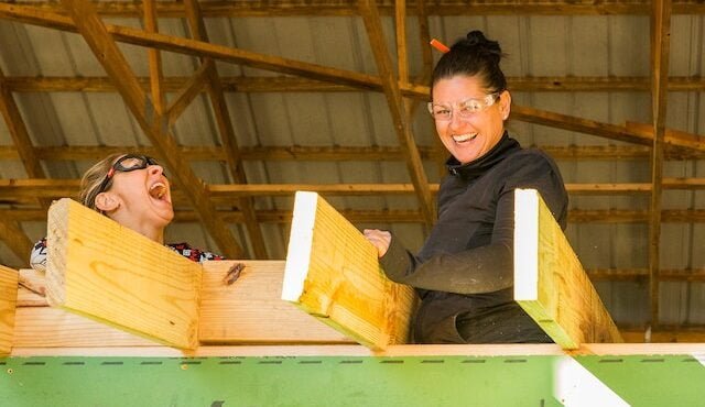 Two women laugh and smile together while building the roof on a tiny home structure in a carpentry workshop