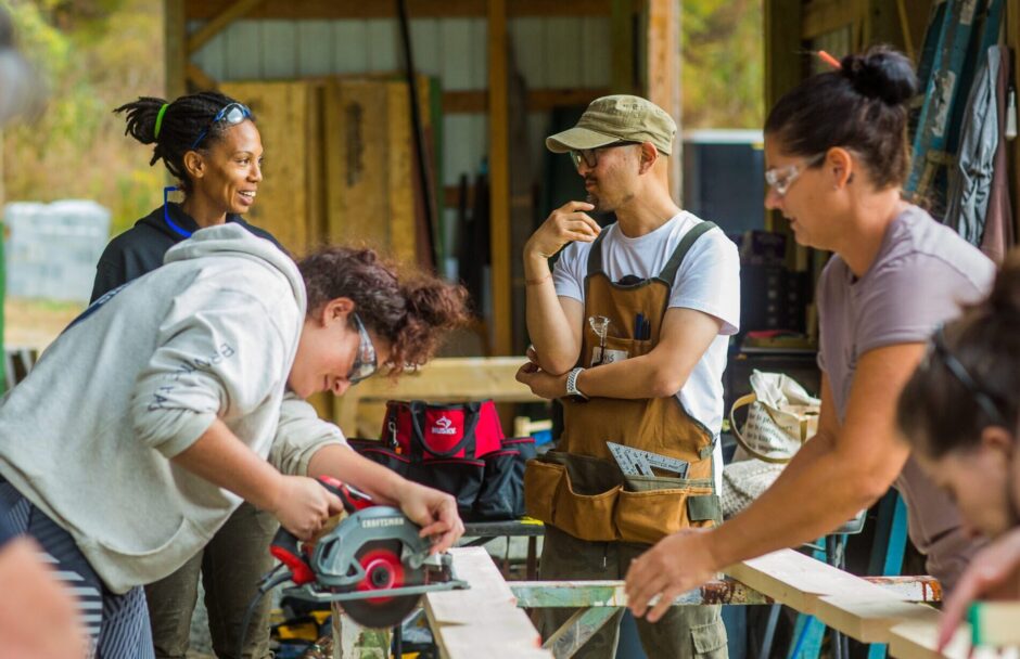 A diverse group of students young and old, male and female take turns practicing sawing wood in a woodworking class