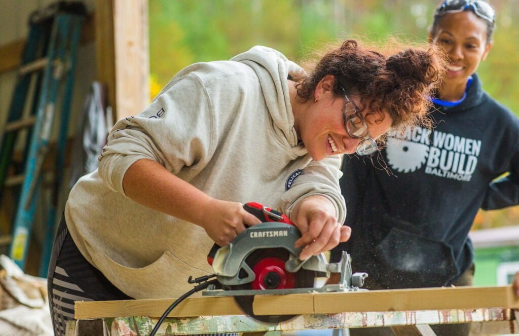 A woman uses a circular saw while another student smiles, watching her success during a tiny house building workshop