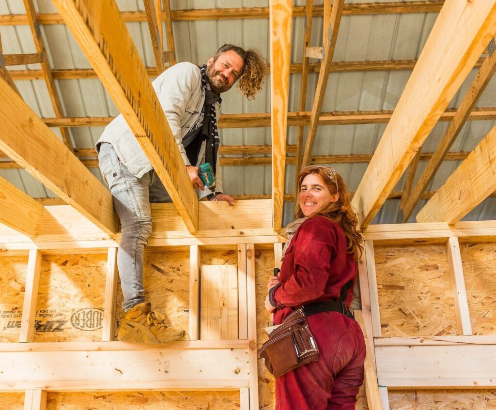 A man and a woman smile while working on the rafter blocking in a tiny house building workshop
