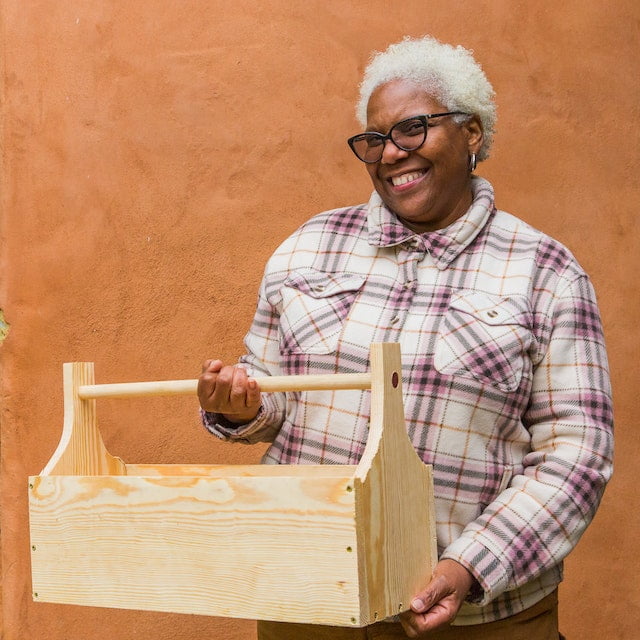 Older black woman smiles triumphantly with the tool caddy she built herself as the final project during a woodworking class for women