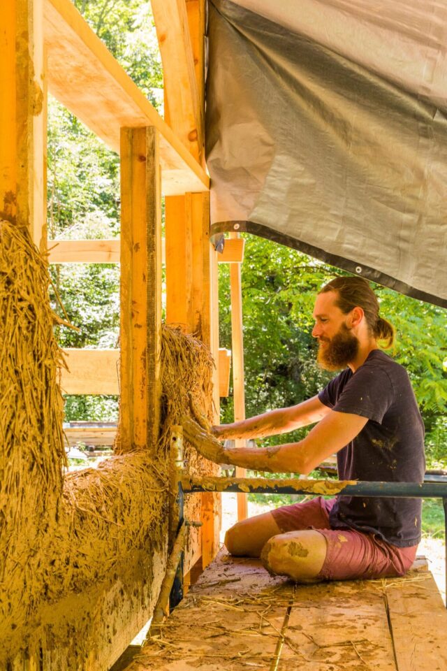 A male student practicing natural building techniques working on a class project building a wall with a mixture of clay, staw, and sand.