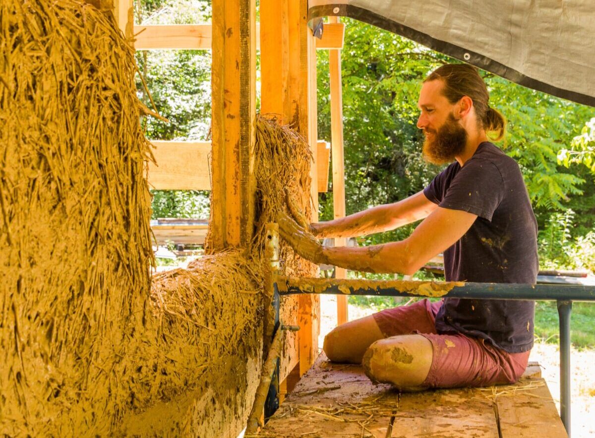 A male student practicing natural building techniques working on a class project building a wall with a mixture of clay, staw, and sand.