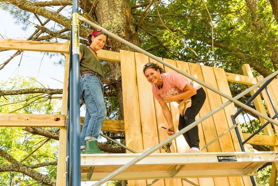 Two women smile down from the worksite of a privacy fence they are learning how to build during basic carpentry class