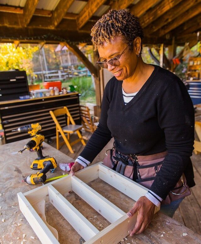 Woman of color smiles down at her carpentry class project: a custom shelf