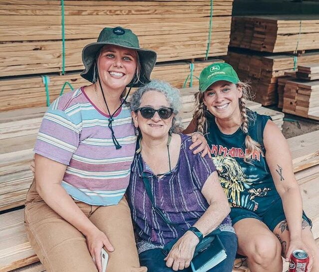 Three women smile and pose with arms around each other at lumber yard