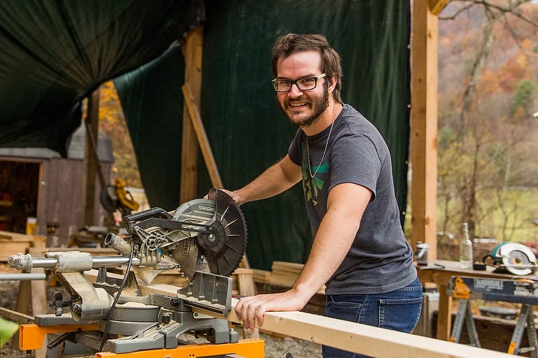 Hayden, a male student, smiles up from preparing to cut a 2x4 on a mitre saw during an adult beginners carpentry workshop at Wild Abundance
