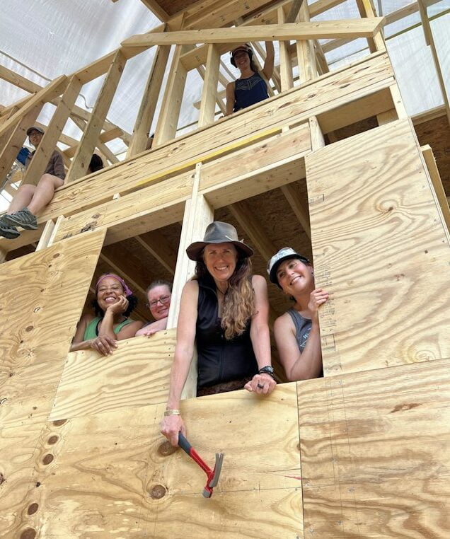 4 students building a tiny house smile as they lean out the windows of the structure they are building for a class photo