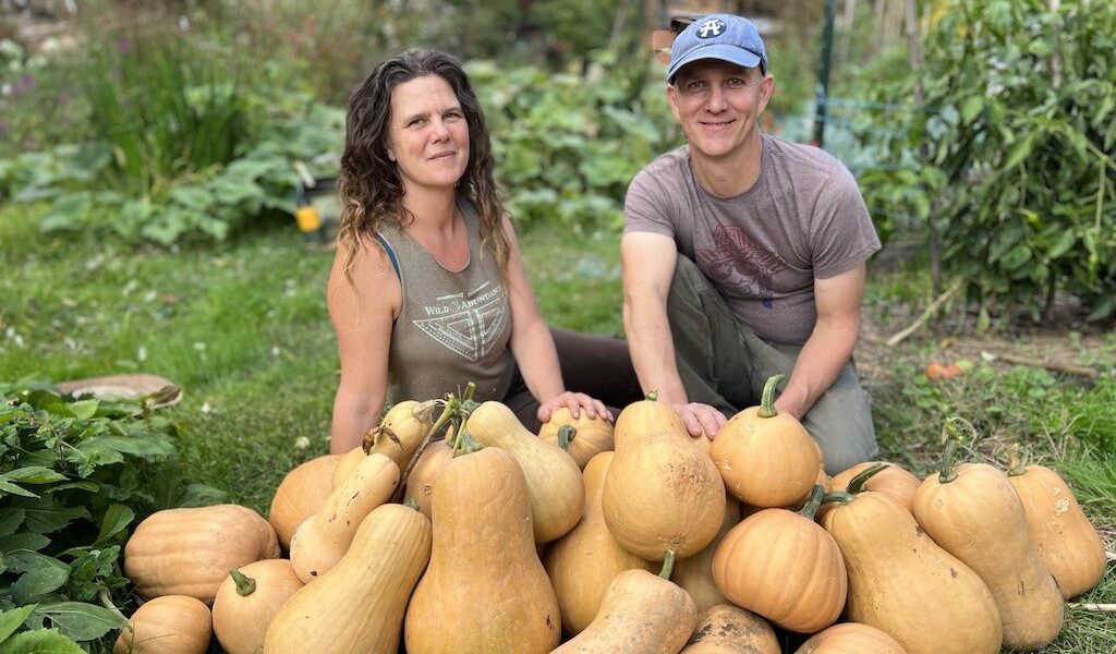 An instructor and a student smile from behind a giant pile of harvested squash at a Gardening and Permaculture Apprenticeship program
