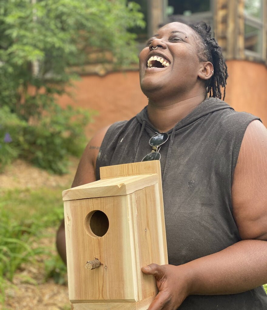 A Black woman smiles holding the birdhouse she built as a final project in the basic carpentry class