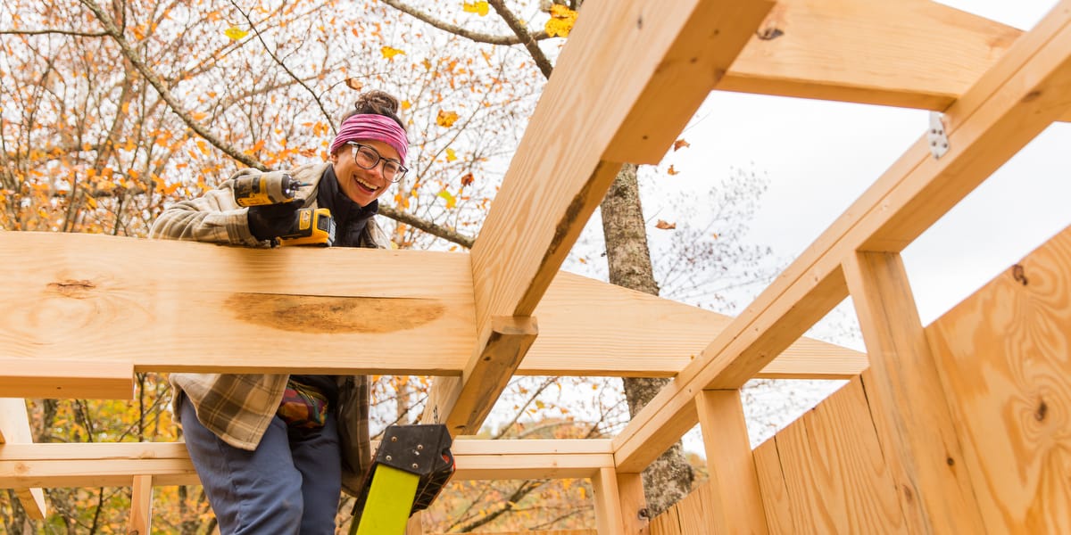 WAC 2025-1200-Current-76 A woman smiles down from fastening rafters together on a small structure she's building with a Women's Advanced Carpentry workshop at Wild Abundance in North Carolina