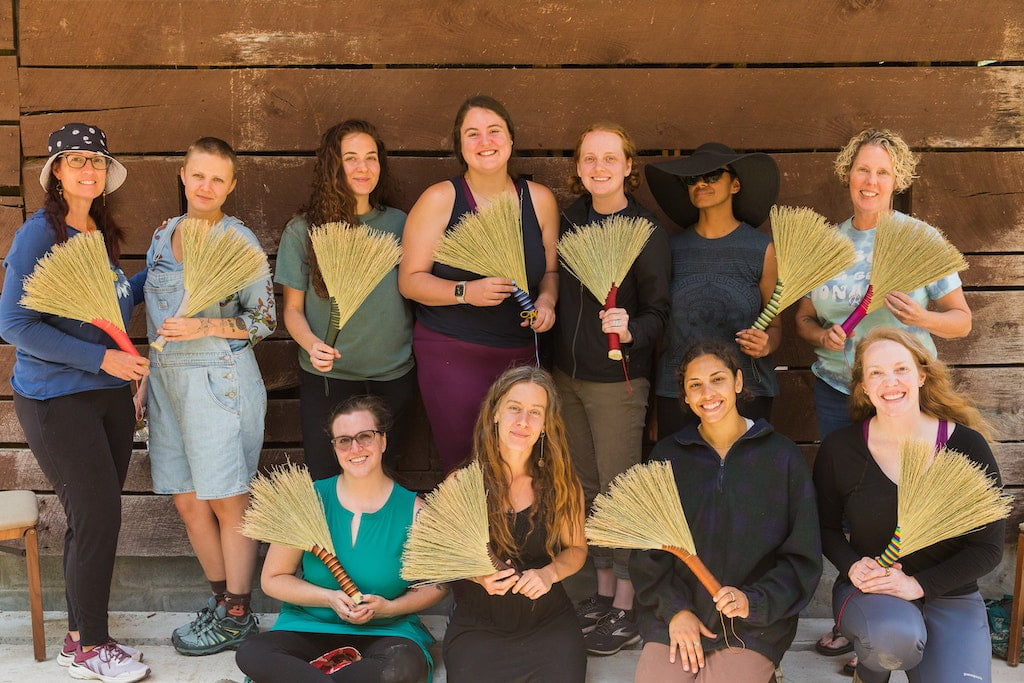 A group of women smile holding their handmade whisk brooms made during a women's Rewilding retreat at Wild Abundance