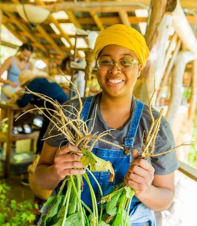 Young black female student smiles while holding up roots she's harvested to make a tincture with at the Wildcrafted Apothecary Medicinal Herbs course at Wild Abundance