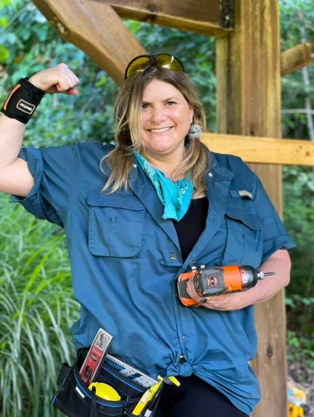 Woman holding a drill and wearing a toolbelt holds up a fist to show strength as she smiles