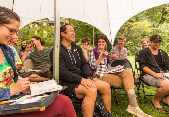 Permaculture Design students smiling and laughing with notebooks out during a PDC class in an outdoor classroom tent