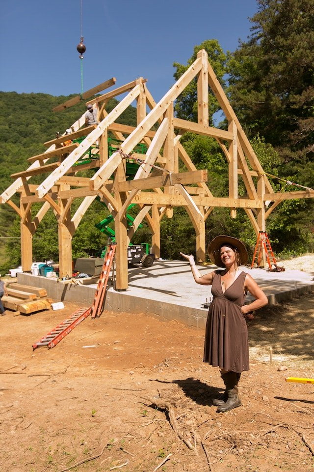 Natalie, the visionary behind Wild Abundance, smiles in front of the Timber Framed Teaching Pavilion mid-construction during a building workshop