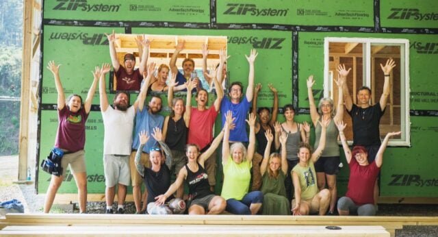  A group of tiny house building workshop students smile and pose with their hands in the air, standing in front of the DIY tiny home they are building in a class