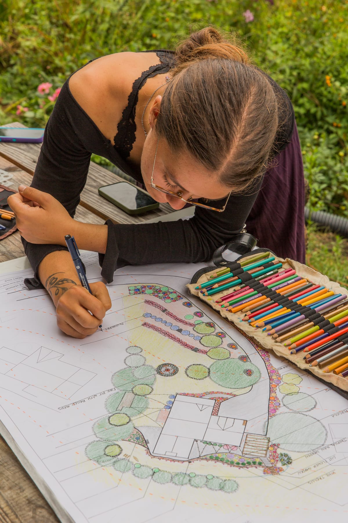 A female permaculture design student shown drawing intricate landscape plans with plant labels and coloring as they prepare their final project to earn their permaculture design certificate at the annual Wild Abundance PDC