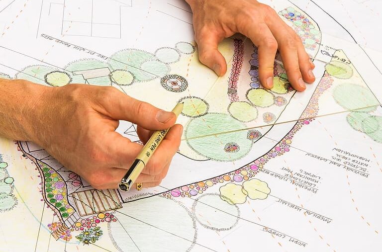 A permaculture design student's hands are shown drawing intricate landscape plans with plant labels and coloring as they prepare their final project to earn their permaculture design certificate at the annual Wild Abundance PDC