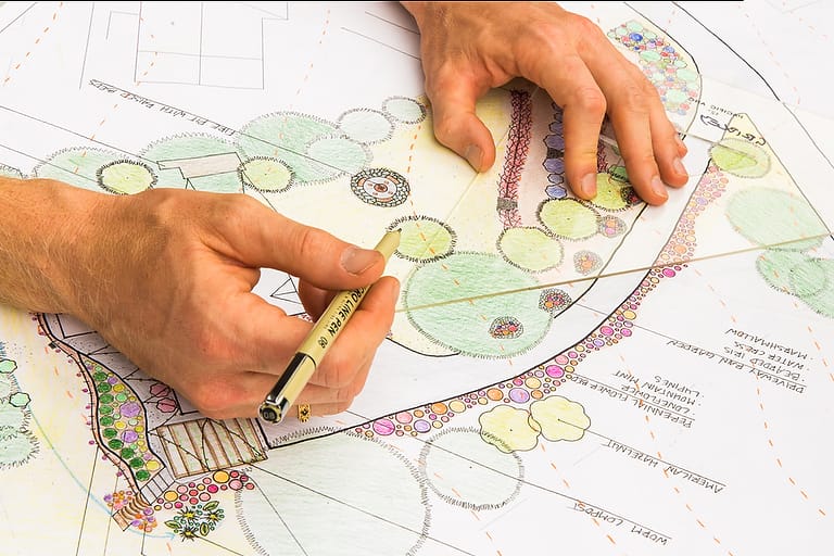 A permaculture design student's hands are shown drawing intricate landscape plans with plant labels and coloring as they prepare their final project to earn their permaculture design certificate at the annual Wild Abundance PDC
