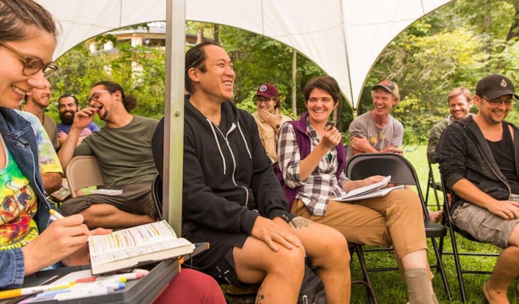 Permaculture Design students smiling and laughing with notebooks out during a PDC class in an outdoor classroom tent