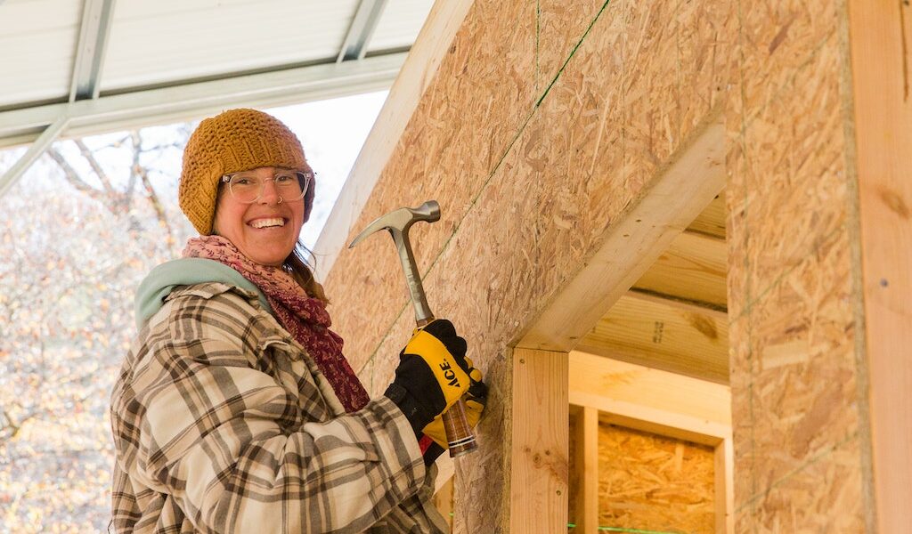 A young woman student smiles down from a ladder where she is working on building a tiny house during a workshop at Wild Abundance.