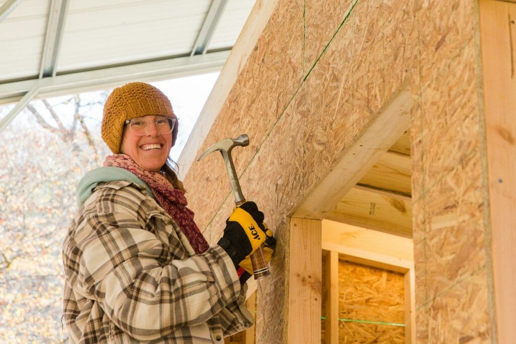 Woman smiles holding a hammer as she nails sheathing to a tiny house in a building workshop at Wild Abundance