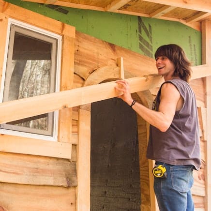 A student in the 4 month carpentry training program at Wild Abundance smiles while lifting a rough cut board up to add siding to a small structure