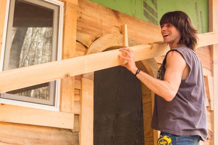 A student in the 4 month carpentry training program at Wild Abundance smiles while lifting a rough cut board up to add siding to a small structure