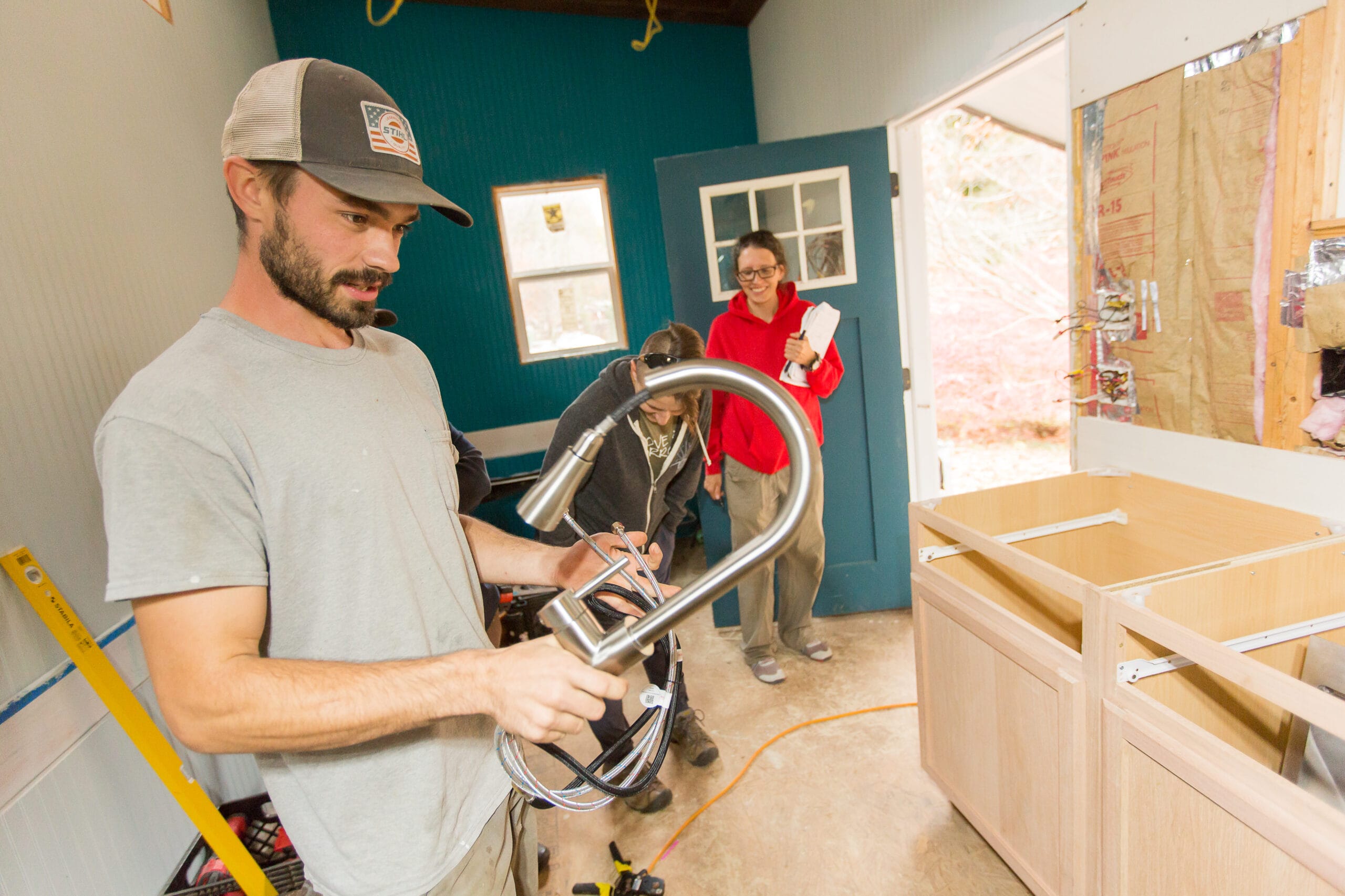 0329-WA-TinyHouseAcademy-©SarahTew-Austin teaching pex plumbing A male Wild Abundance teacher holds a faucet before installing it with students at the Handy Homeowner Bootcamp
