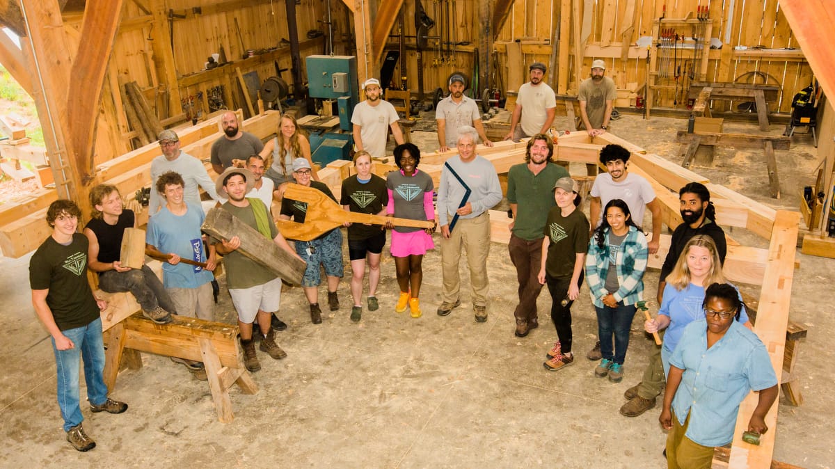 Ivy Creek Timber Framing Workshop Brian Snedeker Campus 2025-1200-Current-648 A group of Timber Framing students smiles up from the workshop they've been learning inside of at Ivy Creek Timer Frames